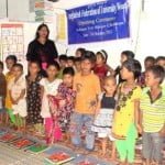 Young children standing in rows in front of a banner with an adult behind them