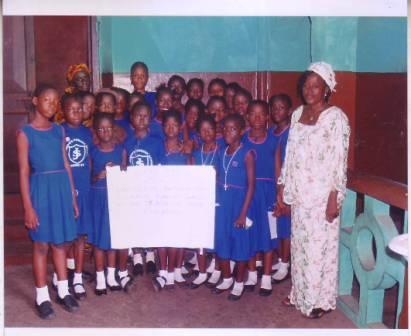 Schoolgirls in blue uniforms standing in a classroom with two women, holding a poster