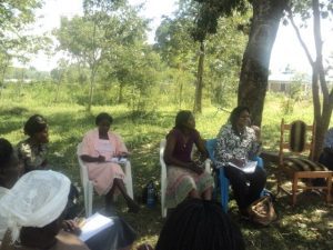 Women seated on chairs outdoors under trees during a project discussion