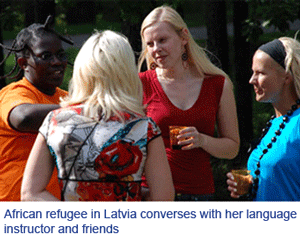 Four women talking together outdoors at a garden gathering. The caption printed on the photograph reads: African refugee in Latvia converses with her language instructor and friends.