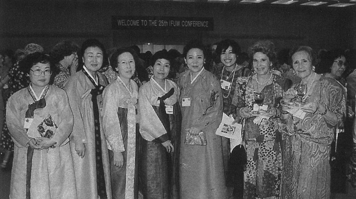 Eight women in traditional and formal dress standing together at the 25th IFUW conference, beneath a welcome banner.