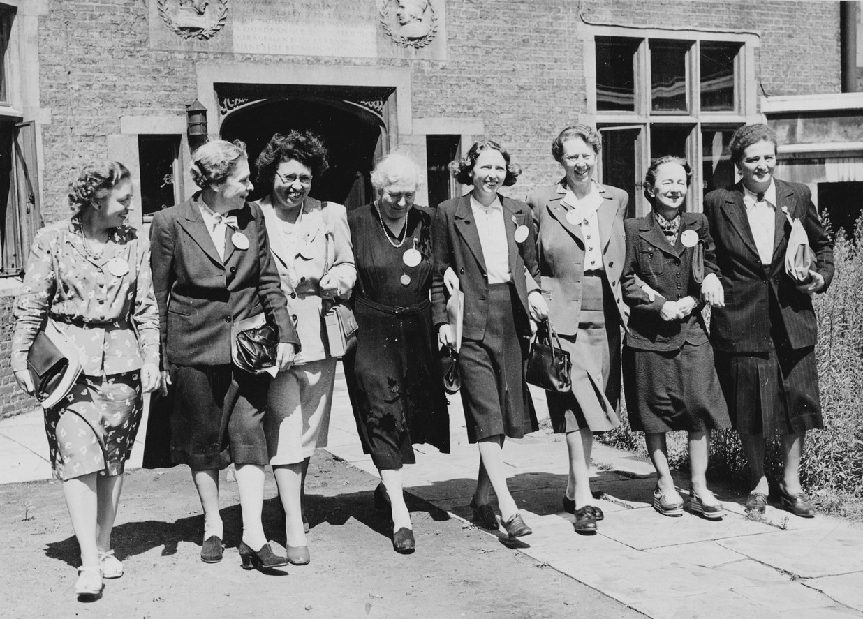 Eight women walking together outdoors, wearing conference badges, in front of a building with a carved stone inscription, 1946.